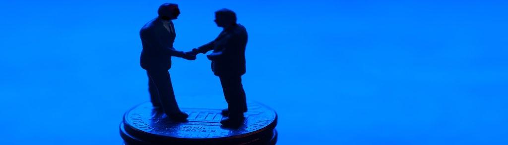 Two miniature figurine businessmen are handshaking on a stack of United States coins. It conceptually represents a financial business deal. Silhouette on blue background.